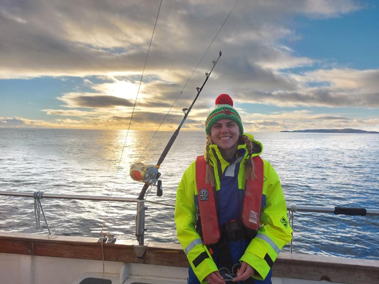 Grace McNicholas, wearing luminescent waterproof gear, smiles on a boat on a calm sea, with a beautiful sky and setting sun in the background. Visible is a heavy-duty rod and reel used to catch tuna, before they are released.