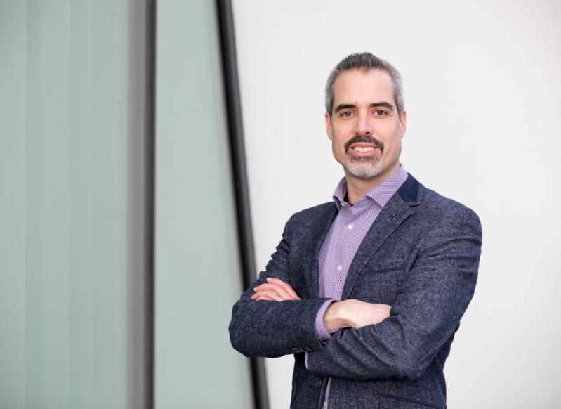 Prof. Kevin Mitchell in a dark blue suit and light purple shirt, smiling outside a Trinity building