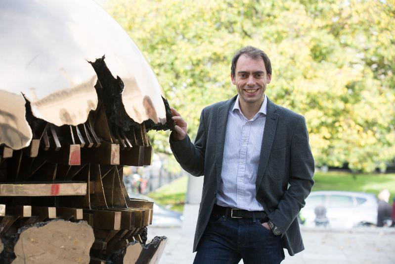 Prof Marco Ruffini, wearing a suit and light shirt, standing next to Trinity's golden sphere sculpture, The Pomodoro, on a sunny day.