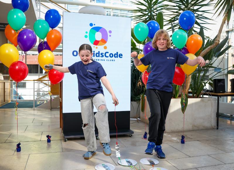 Two children jump in front of an OurKidsCode project poster, with bright balloons to their side.