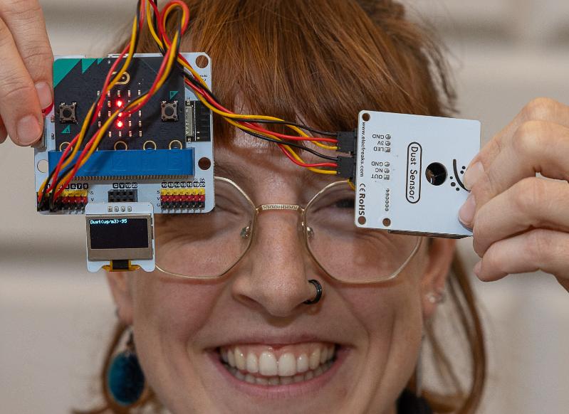 A woman with red hair holds up a circuit board and an electronic plate with wires