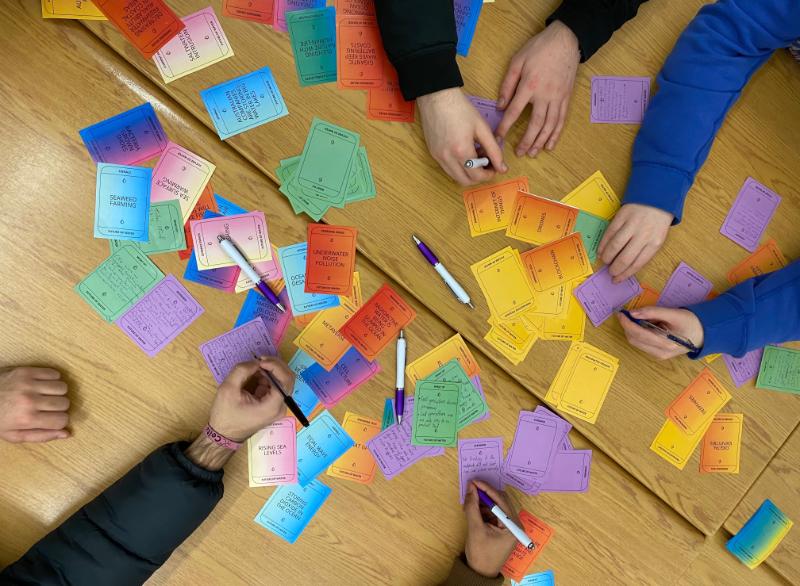 As seen from above, hands reach into the centre of a desk, which is covered in colourful sticky notes.