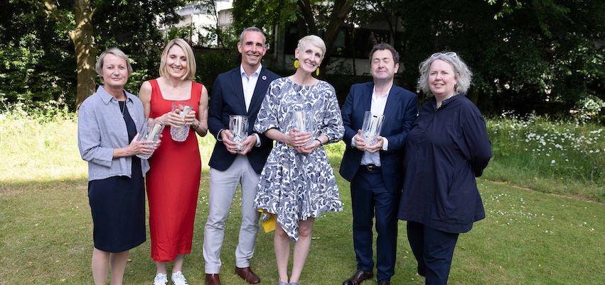 Six people standing in a garden. They are smiling at the camera and five of them are holding awards